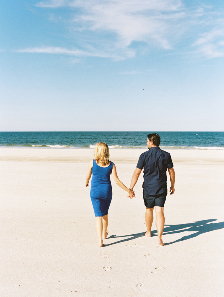 a couple holding hands on a sunny summer day walking away from the camera toward the ocean