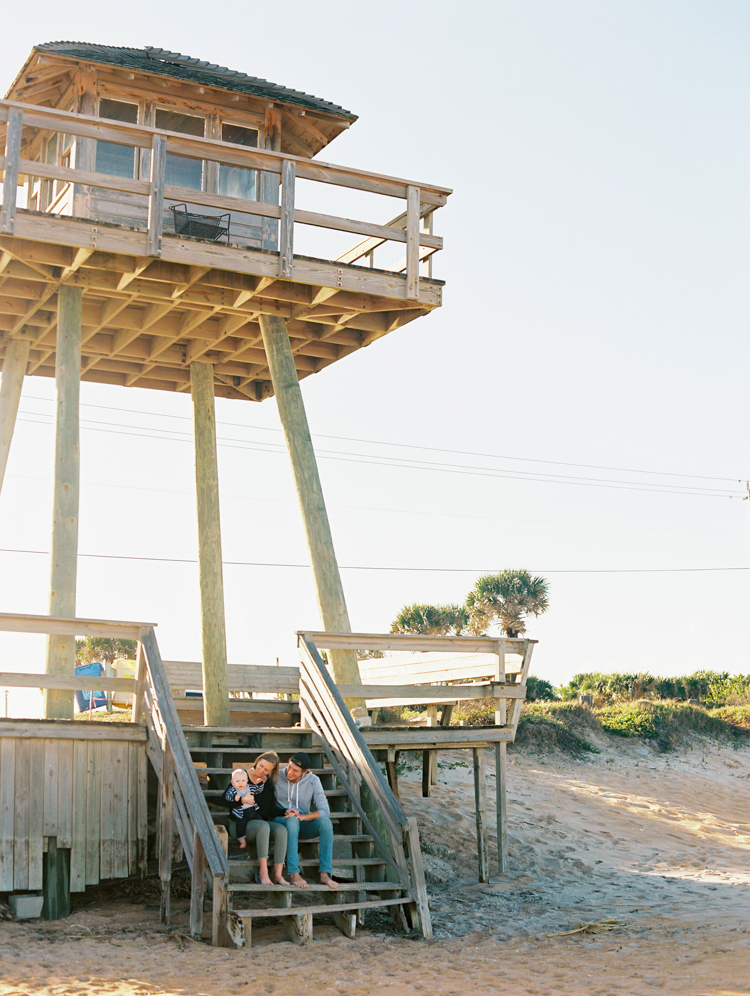 Family of three sitting on the steps of a wooden beach watchtower in the sun.