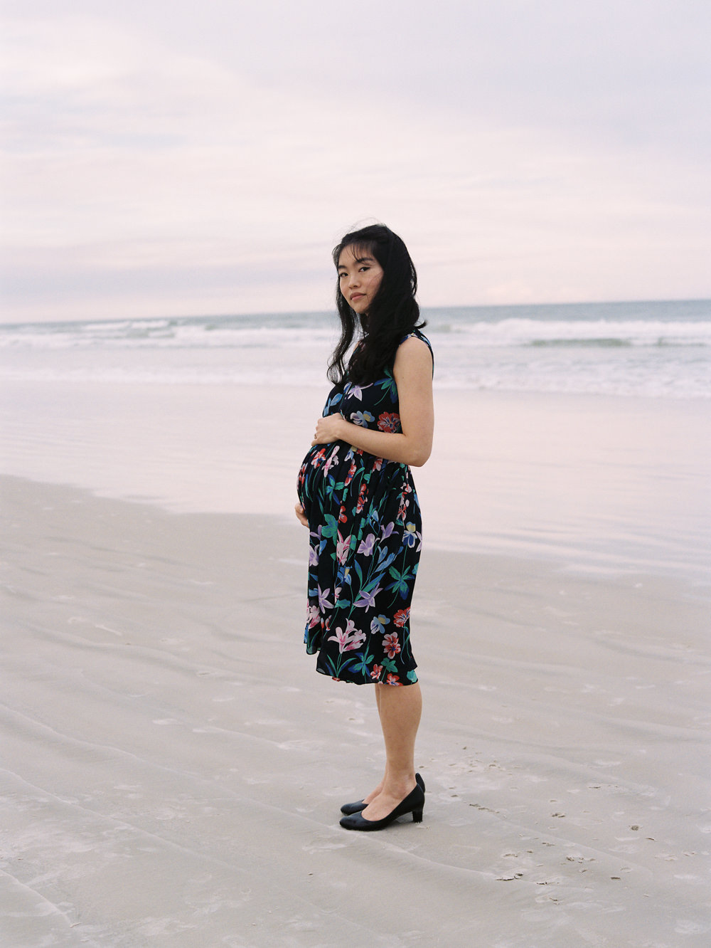 black haired pregnant woman standing on the beach with hand on belly