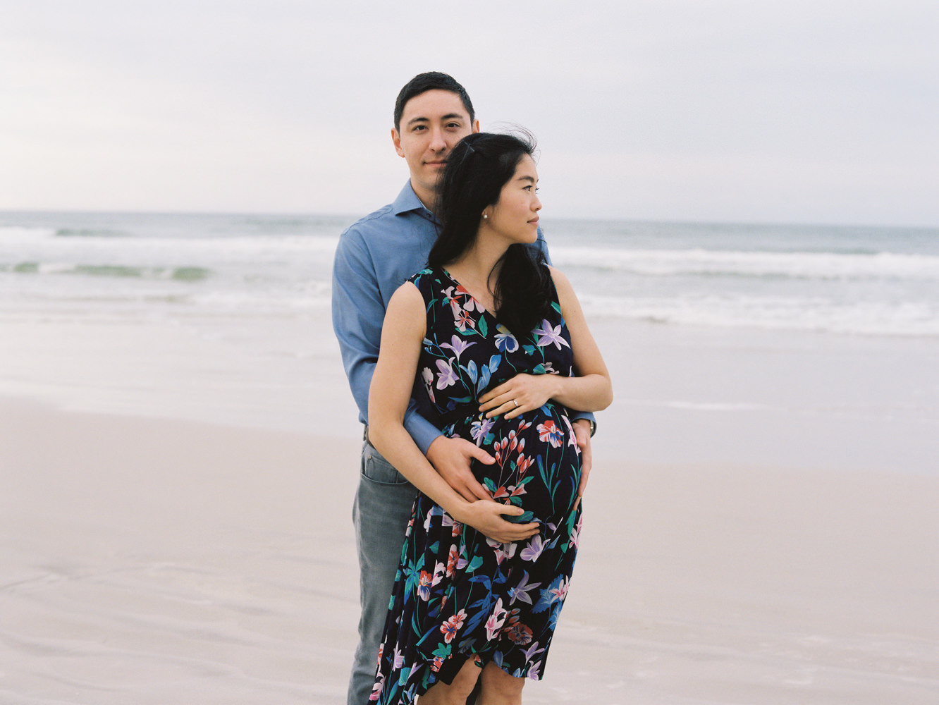 man hugging his pregnant wife from behind and looking calmly at camera while she looks to the side on the beach