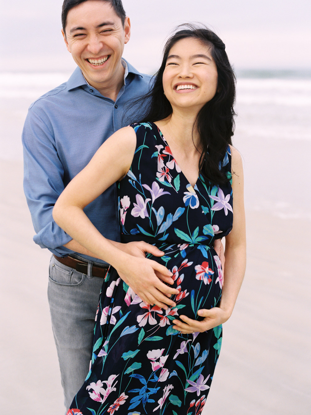 man standing behind woman with his hands on her pregnant belly as they smile and laugh