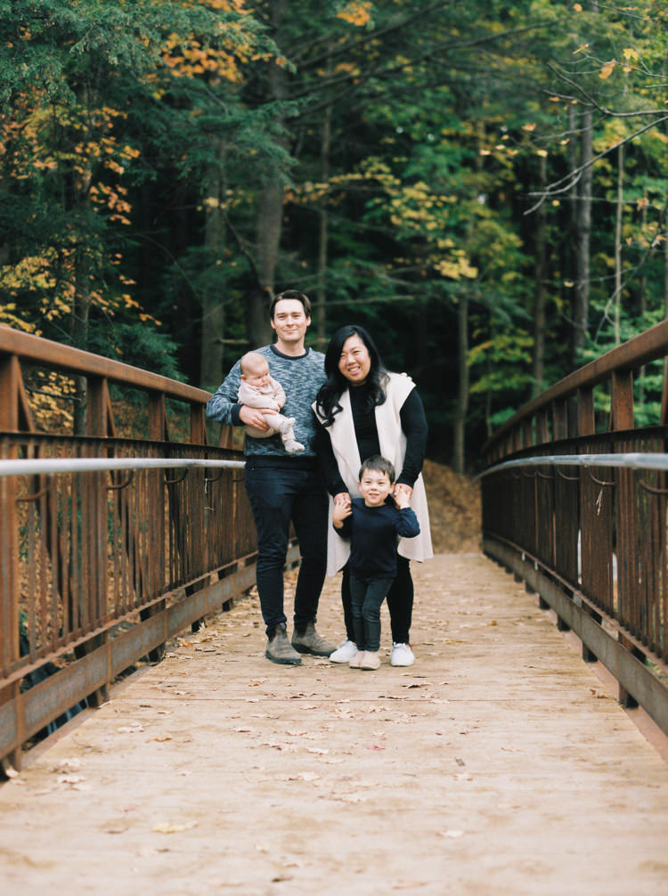 family of four standing on a bridge in a fall forest smiling at the camera