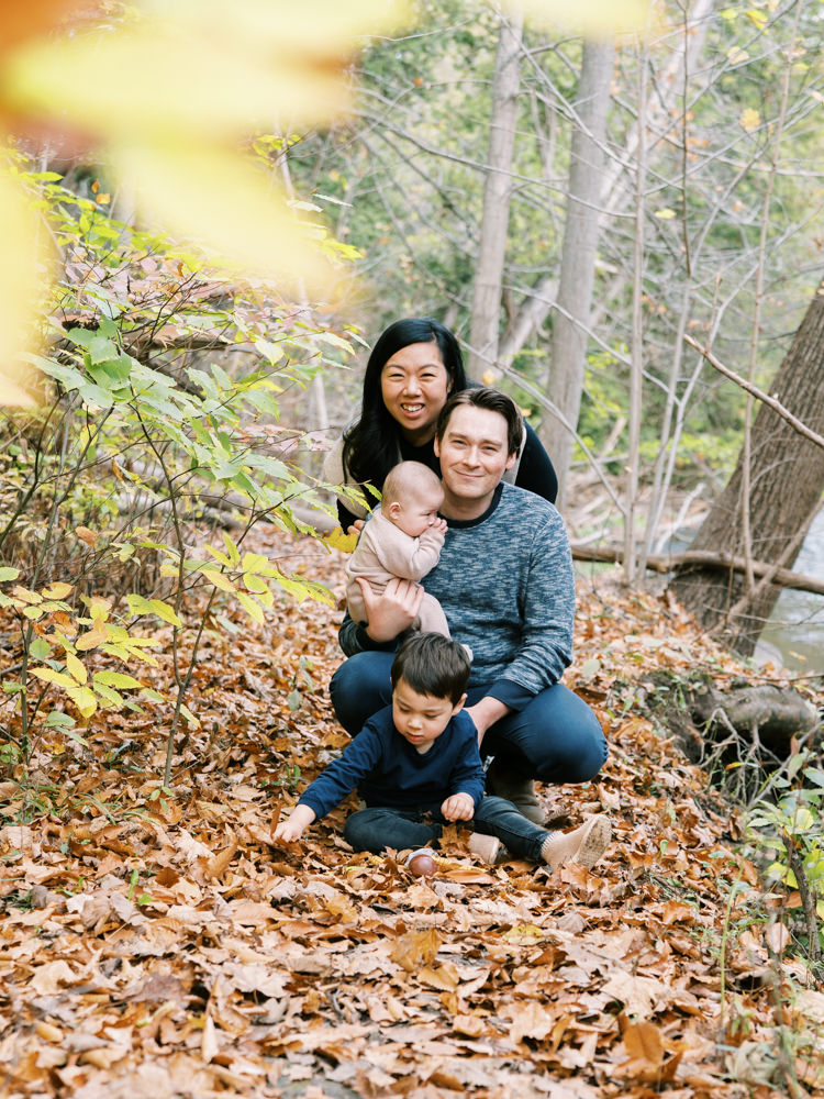 family of four sitting in fall leaves in the woods and smiling at the camera