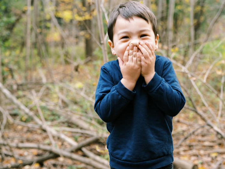 toddler boy in navy blue shirt with hands over mouth laughing at the camera