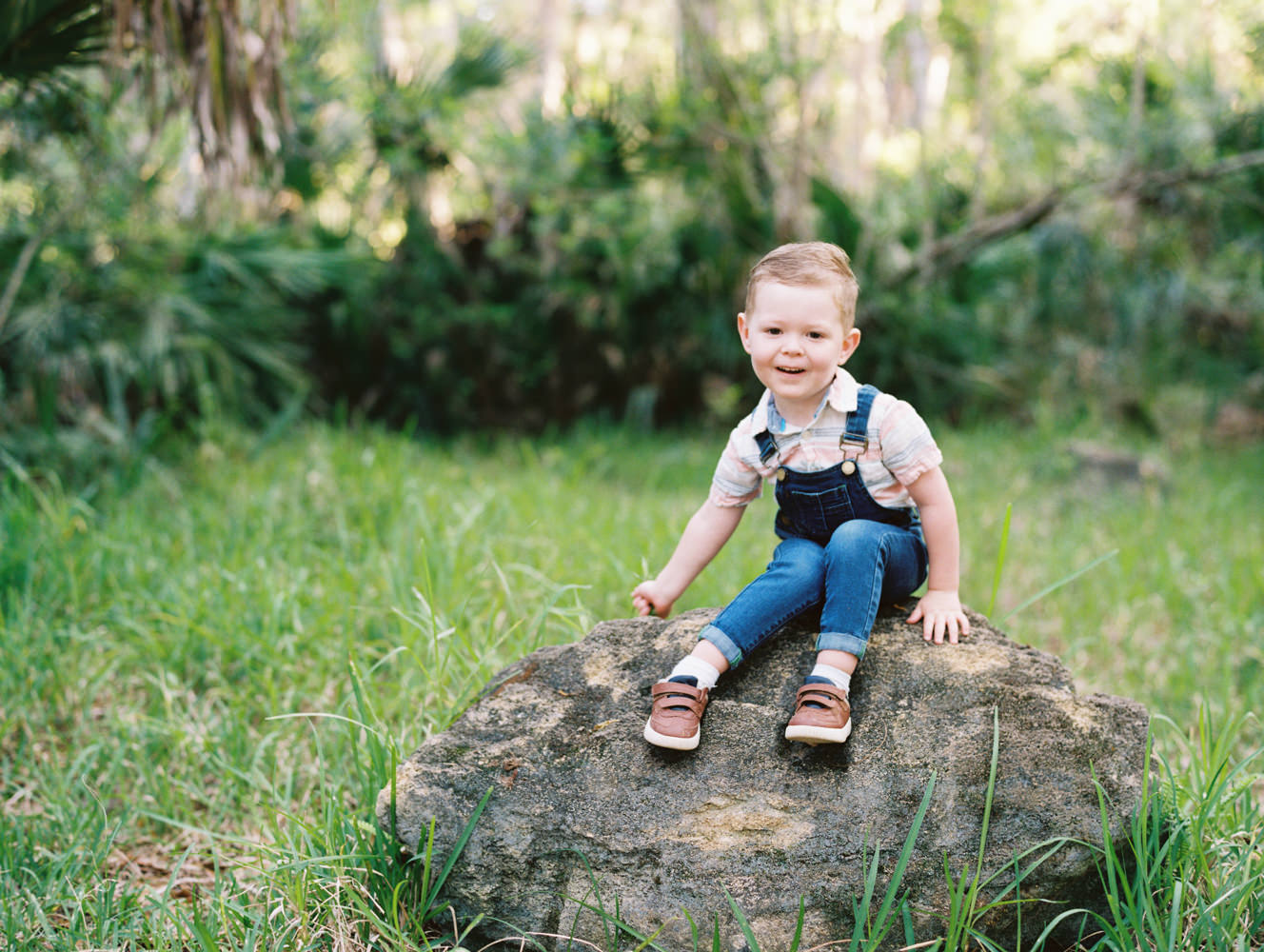 red haired boy wearing overalls sitting on a rock and picking grass