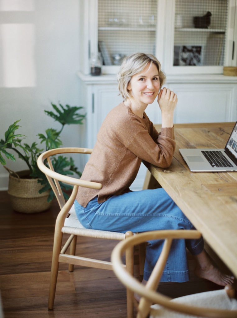 Woman in a brown sweater and jeans sitting a large wood kitchen table in a brightly lit home