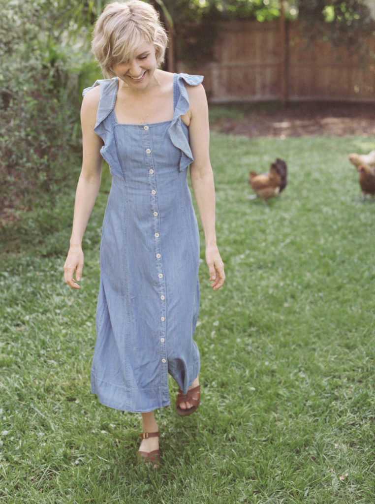 Woman with short blonde hair smiling in a blue dress walking through a yard with chickens 