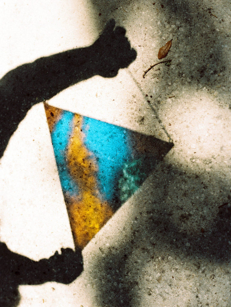 Shadow and reflection on the concrete sidewalk of a person’s hands holding a blue and yellow triangle hanging stained glass piece