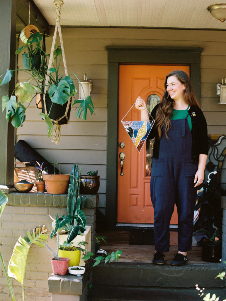 Female stained glass artist standing on her plant filled front porch in navy blue cotton overalls holding blue and yellow stained glass art for brand photos