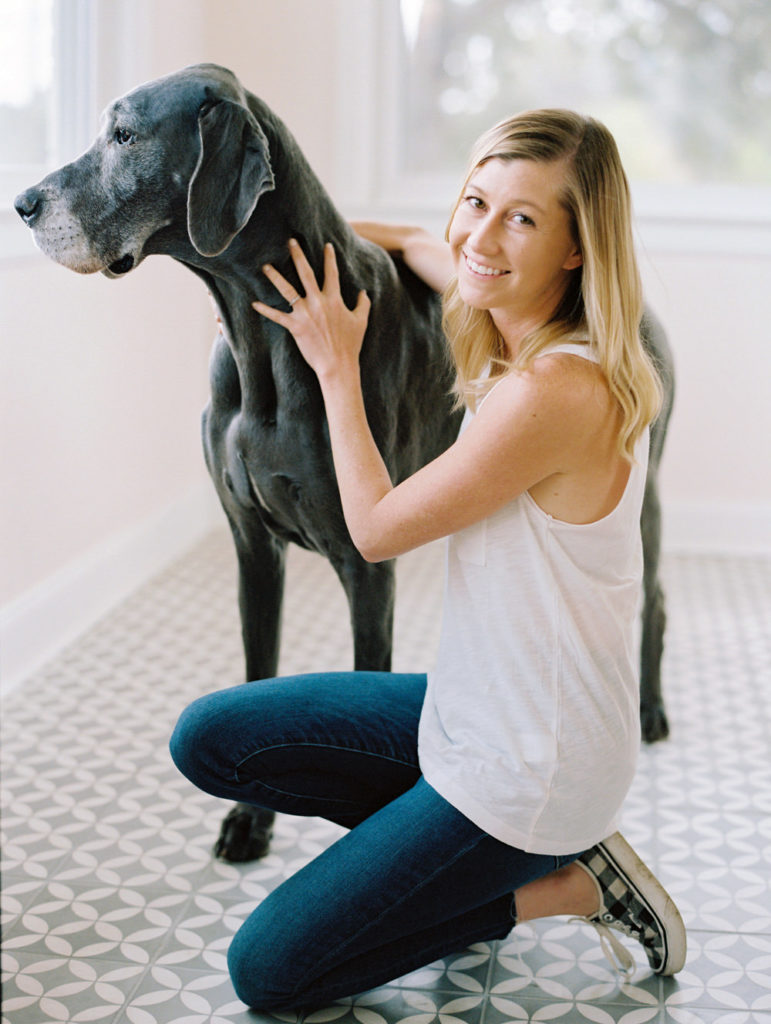 White woman with shoulder length blonde hair wearing a white tank top and jeans, kneeling down next to her grey great dane in a white living room