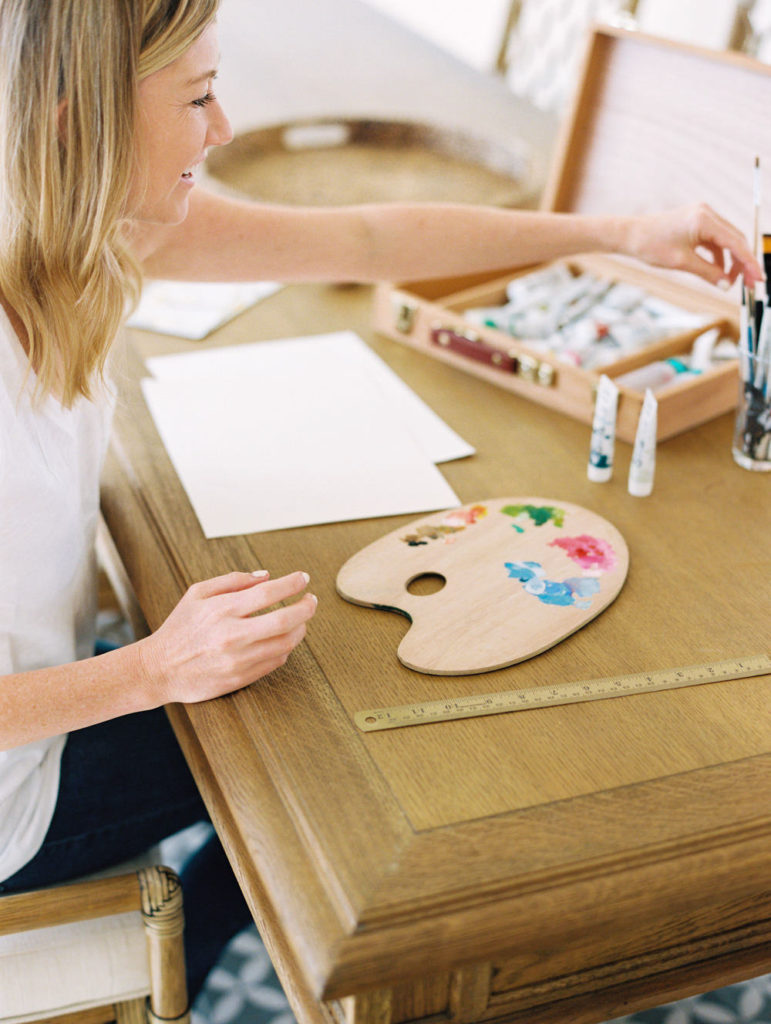 White female artist with blonde hair sitting at her wood dining table setting up art supplies for acrylic painting