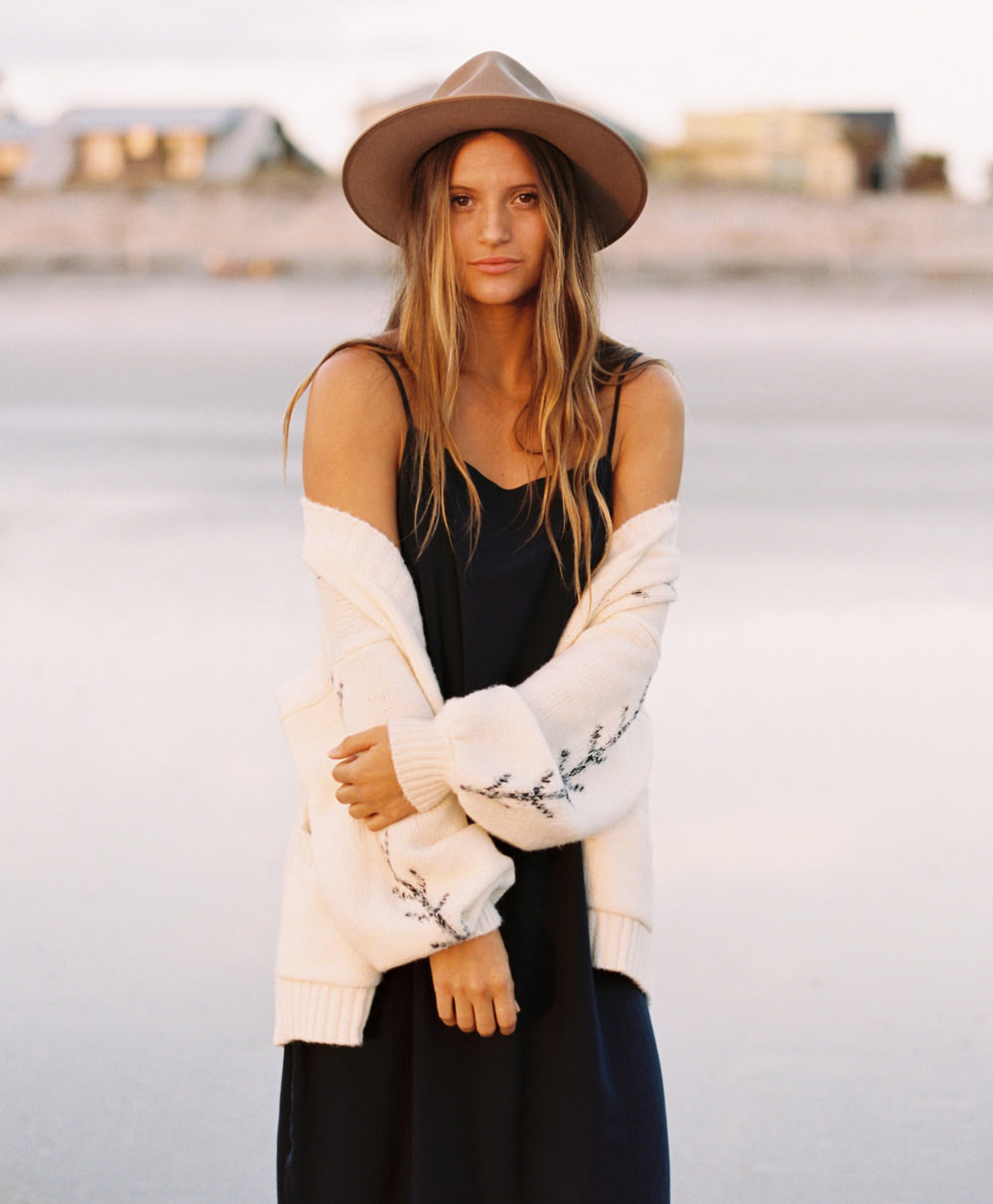 Tan woman with long light brown hair and brown eyes, wearing a brown felt hat, navy blue dress and white sweater staring straight at the camera