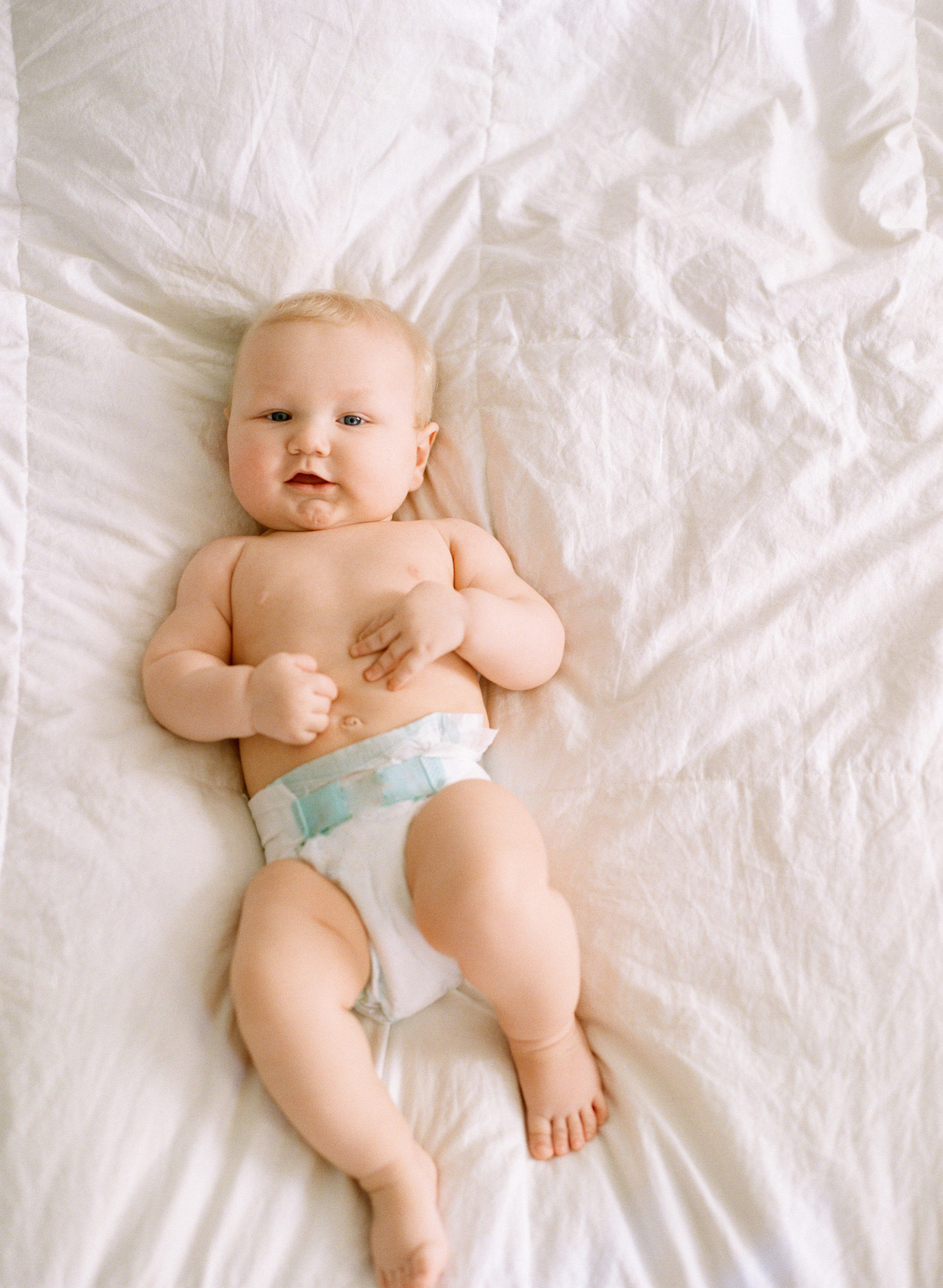 chubby six month blonde baby on a white duvet looking at the camera