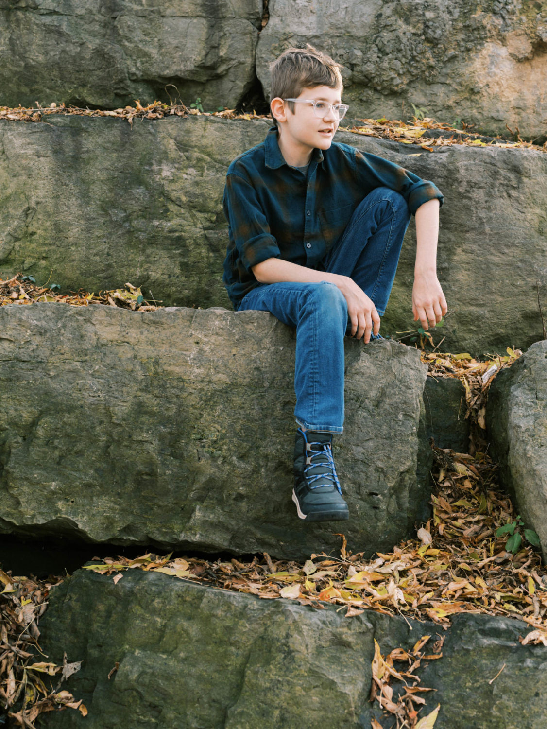 young teen boy in glasses and blue shirt and pants sitting on a boulder at a park in Mississauga