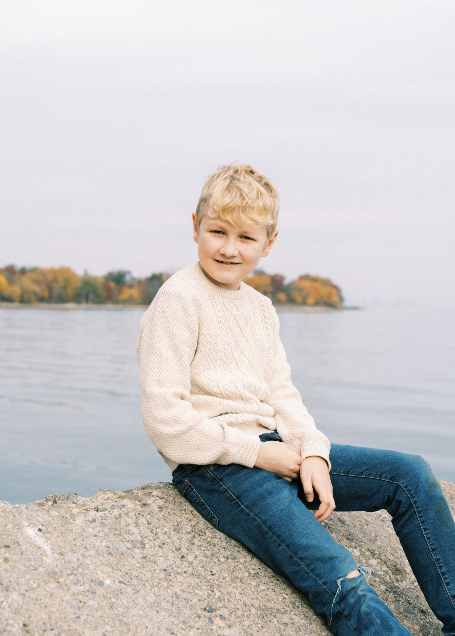 blonde boy in white sweater and jeans sitting on a rock by lake ontario