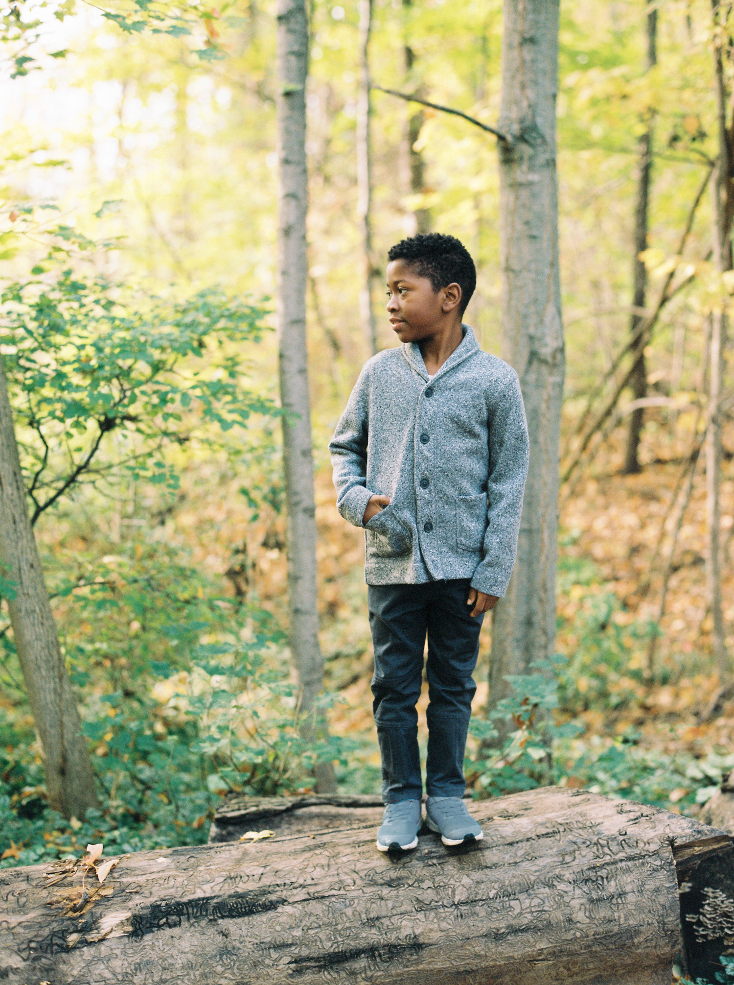 black boy wearing gray sweater and black pants standing on a fallen tree in the forest.