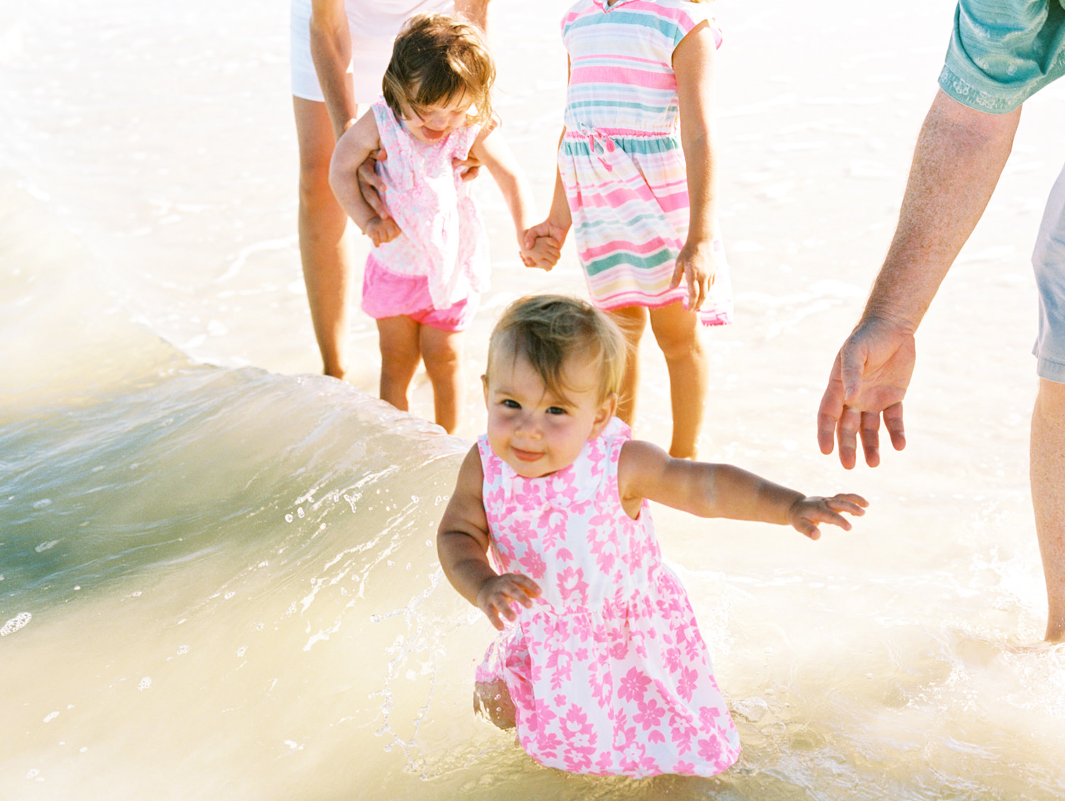 three girls in pink dresses playing in the water while mom and grandpa hold their hands