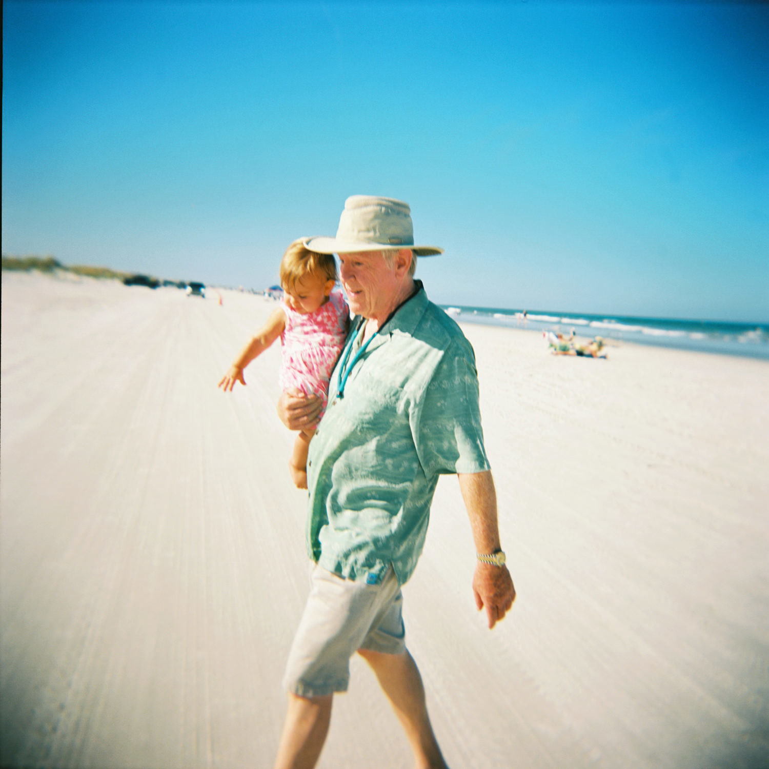 grandfather in hat green shirt and shorts holding young grandchild and walking across beach
