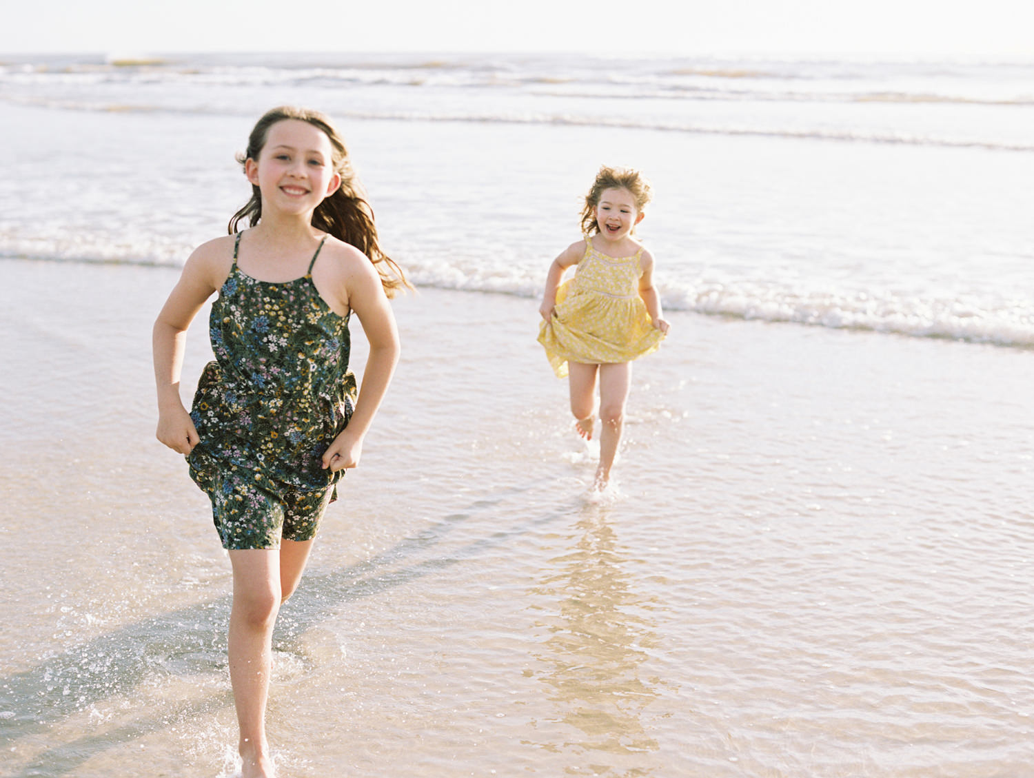 Two sisters in green and yellow dresses running away from waves at the ocean