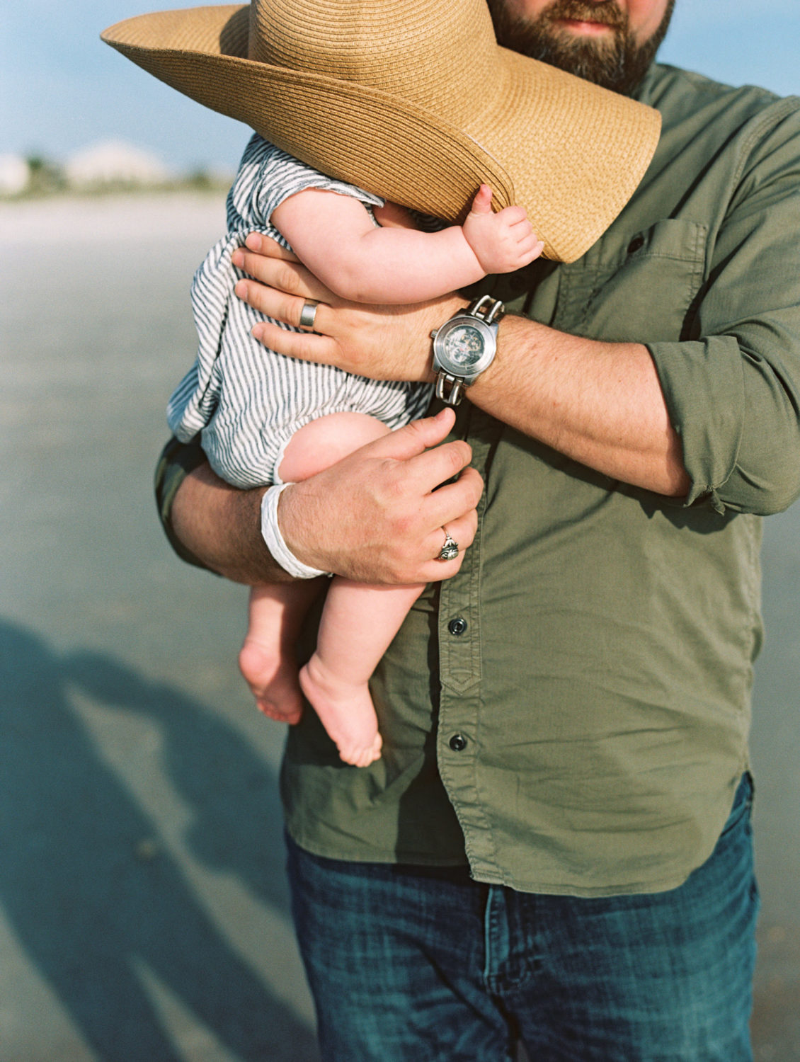baby girl in striped romper wearing mom's sunhat while dad in green shirt holds her.