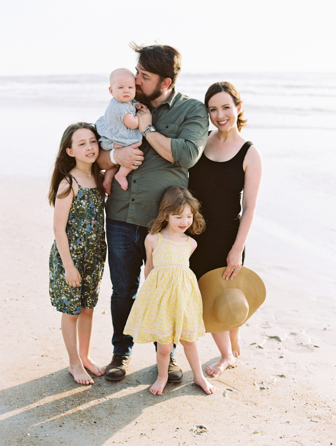 family of five standing close together with bare feet on a sandy beach.
