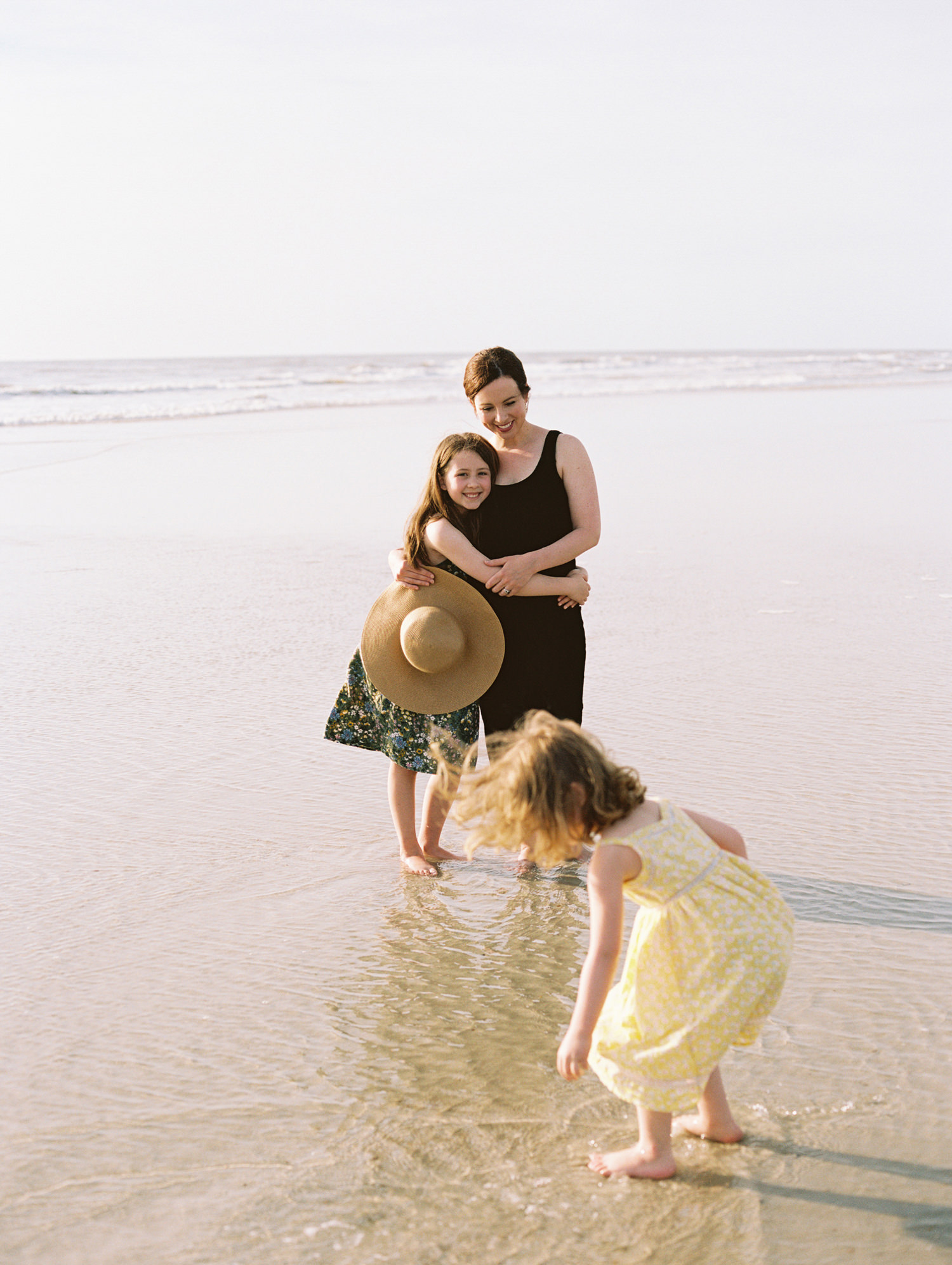 mom in black dress at the beach, hugging older daughter while younger child picks up seashells