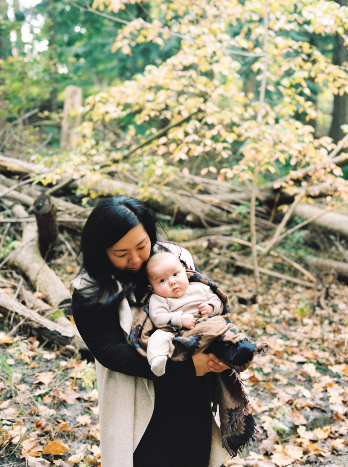 mom with long black hair wearing black and tan sweater standing in autumn woods and smiling down and the baby she's holding