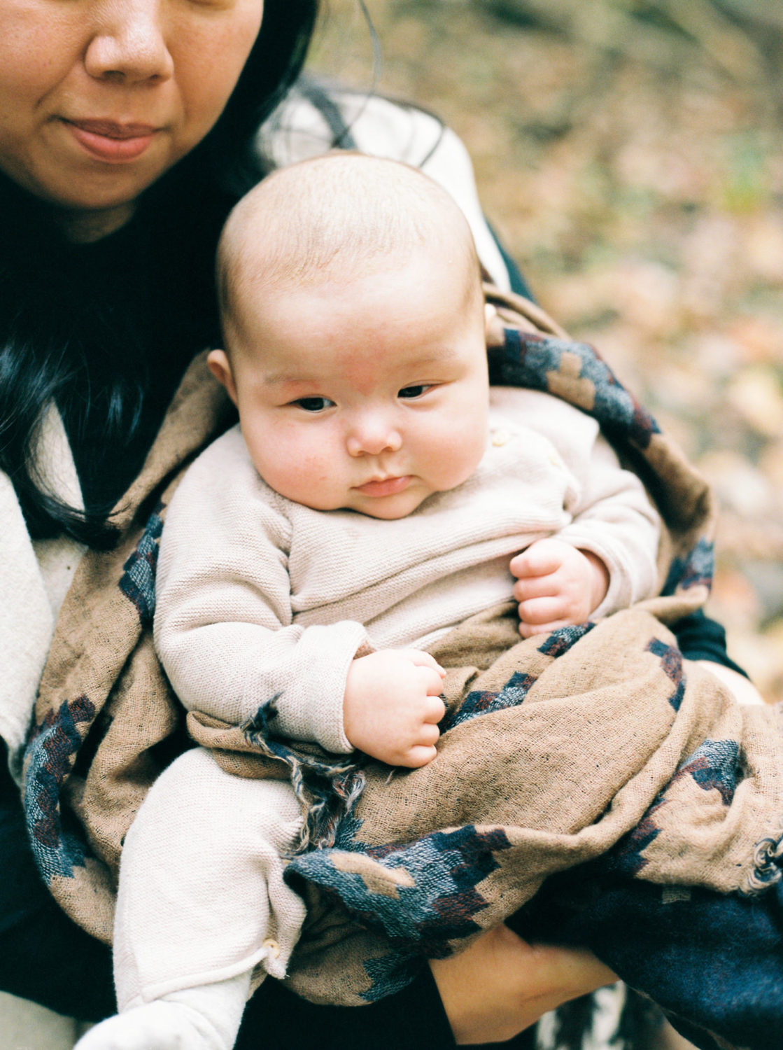 baby with round rosy cheeks in tan jumper wrapped in brown and gray blanket and held closely by mom