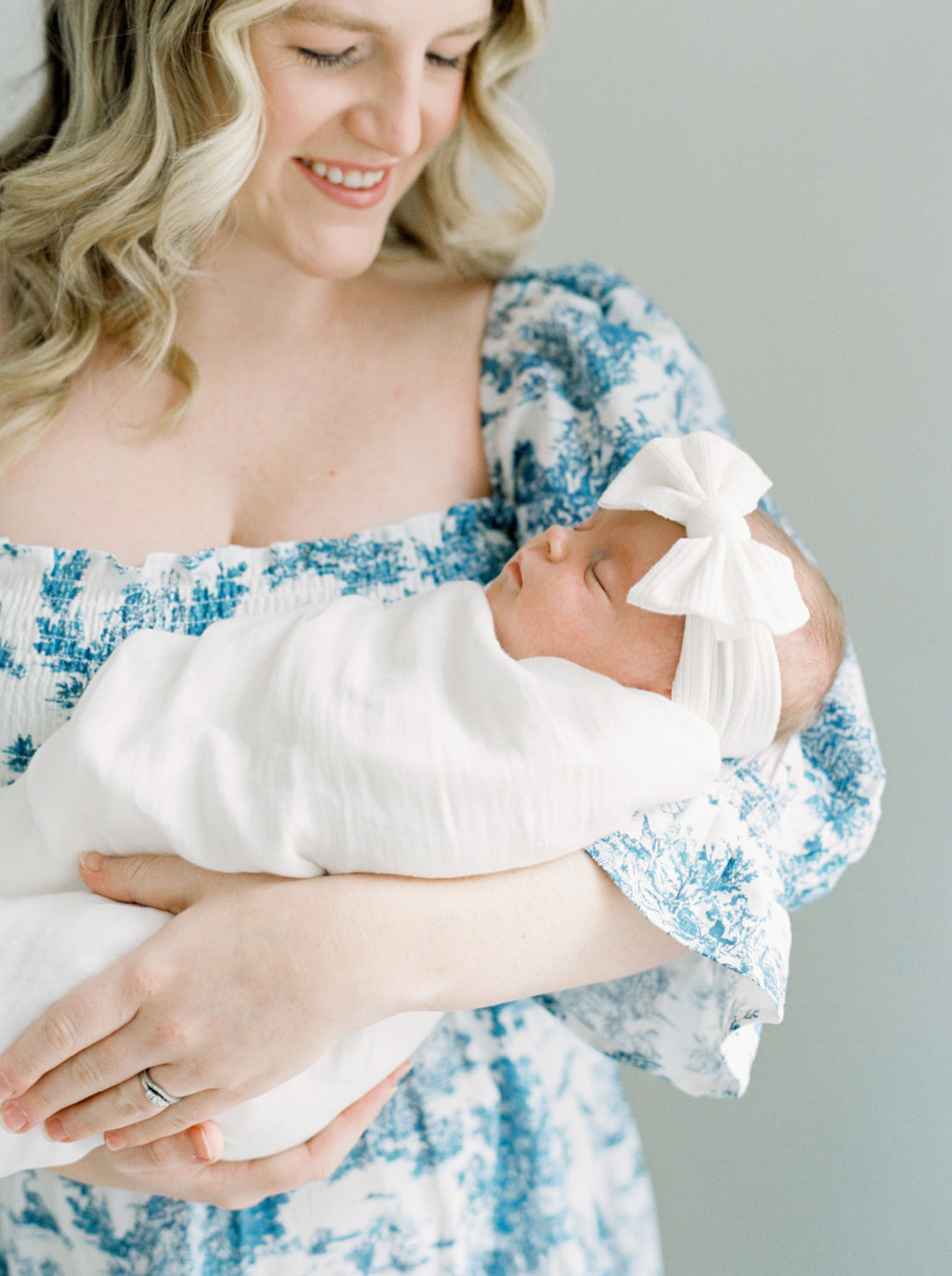 smiling woman with blonde hair in blue and white dress holding a sleeping baby wrapped in white blanket wearing a white bow