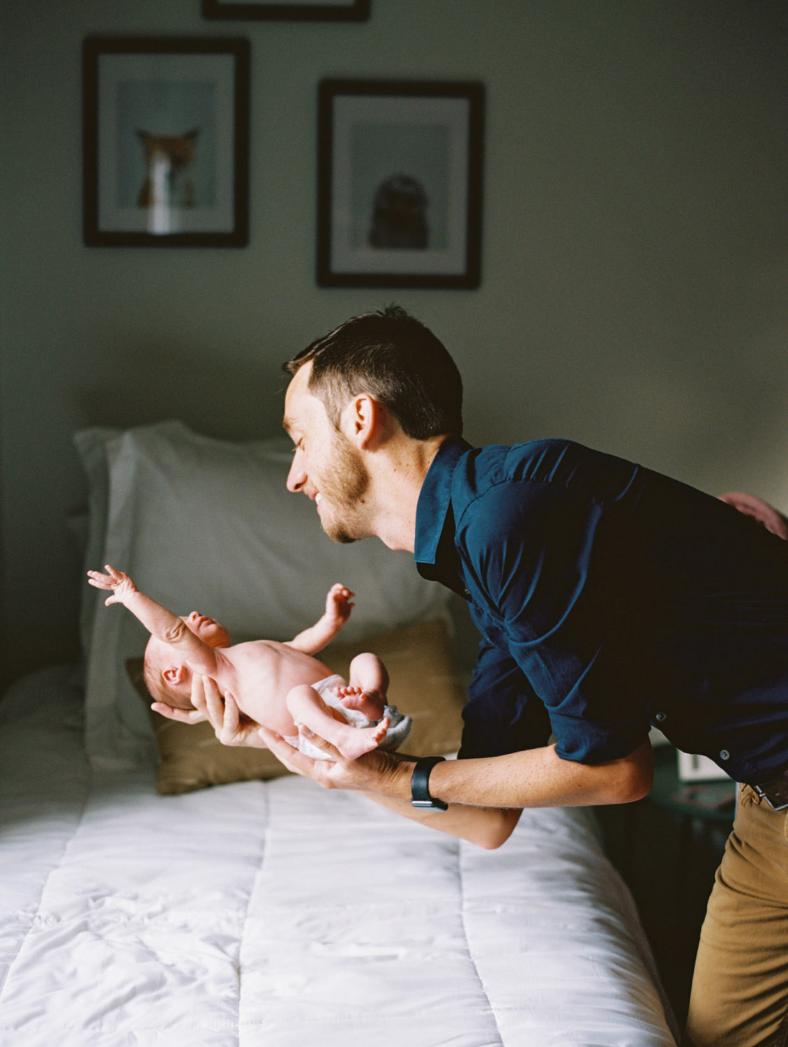 dad in dark blue shirt and tan pants smiling and gently laying newborn stretching its arms on a bed with white blanket.