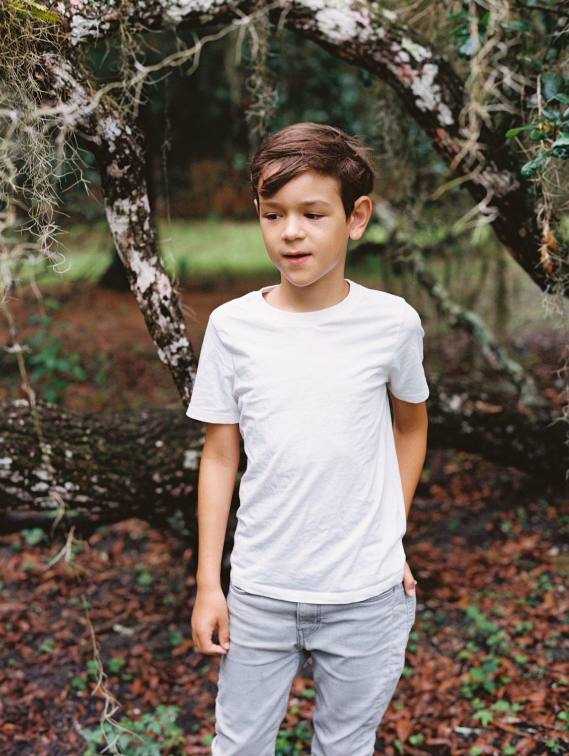 boy with brown hair in white tshirt and gray pants standing in a forest looking to the left