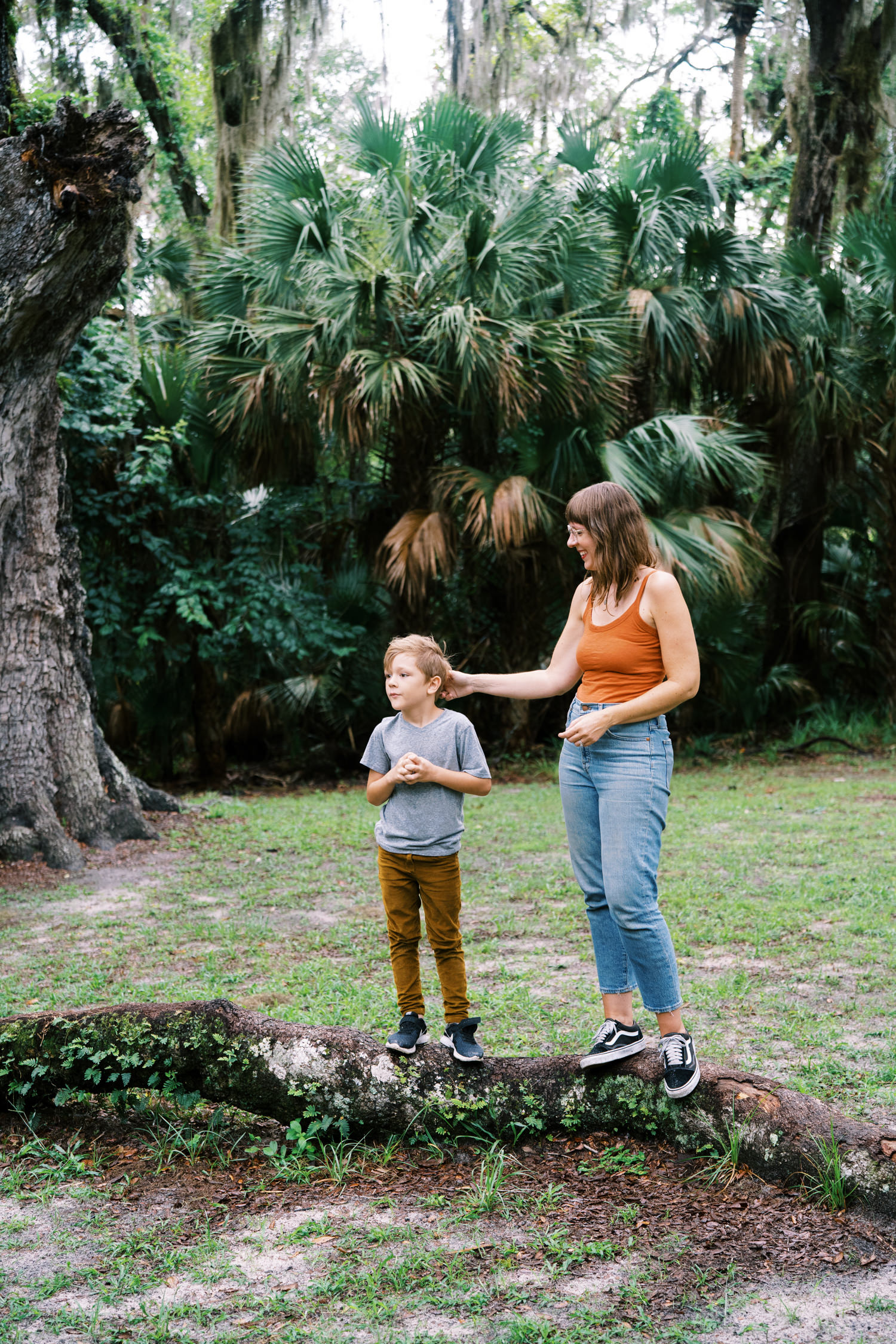 woman in rust color tank top and jeans standing next to her young son stroking his hair and smiling