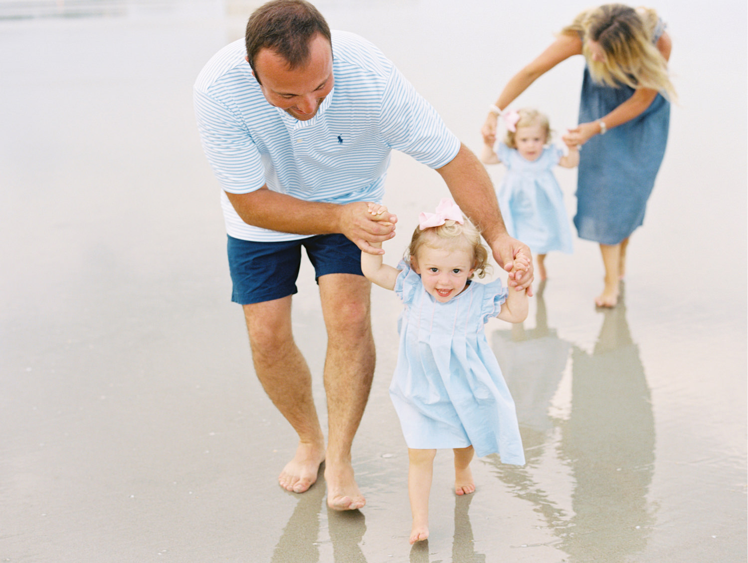 dad in blue shirt and shorts holding his toddler daughters hands and helping her walk on the sandy beach