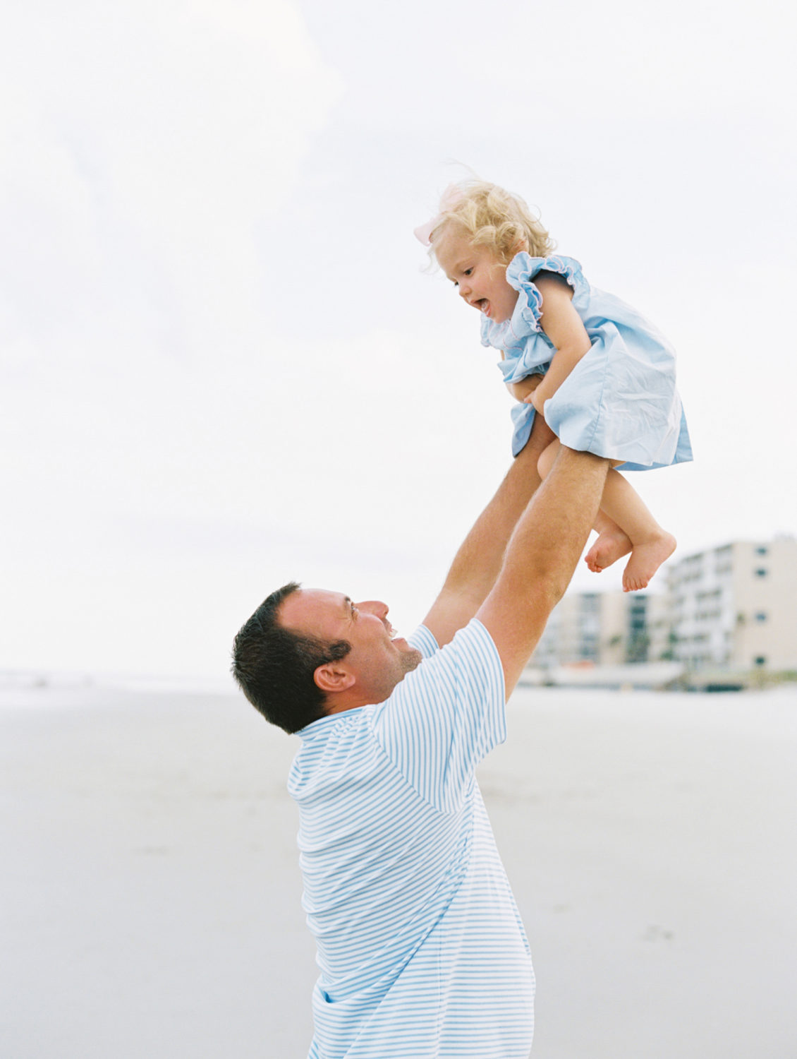 dad in blue shirt lifting toddler in blue dress into the air and both laughing