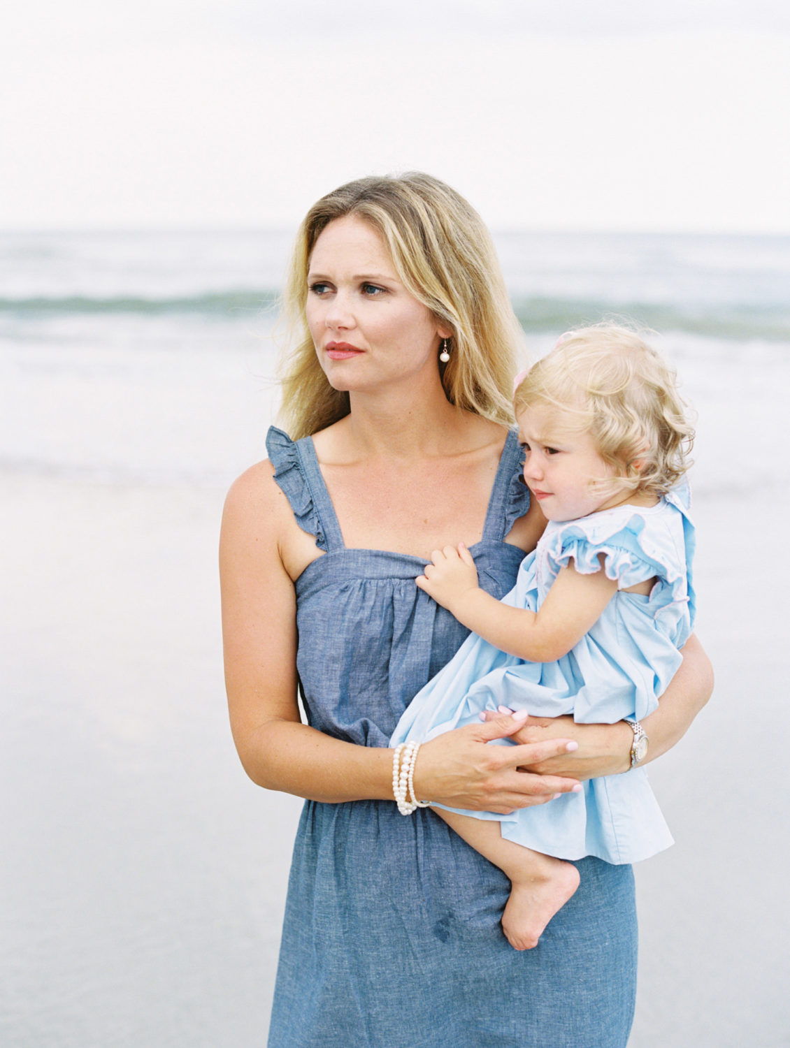blonde woman in blue chambray dress holding toddler in blue dress and looking to the left