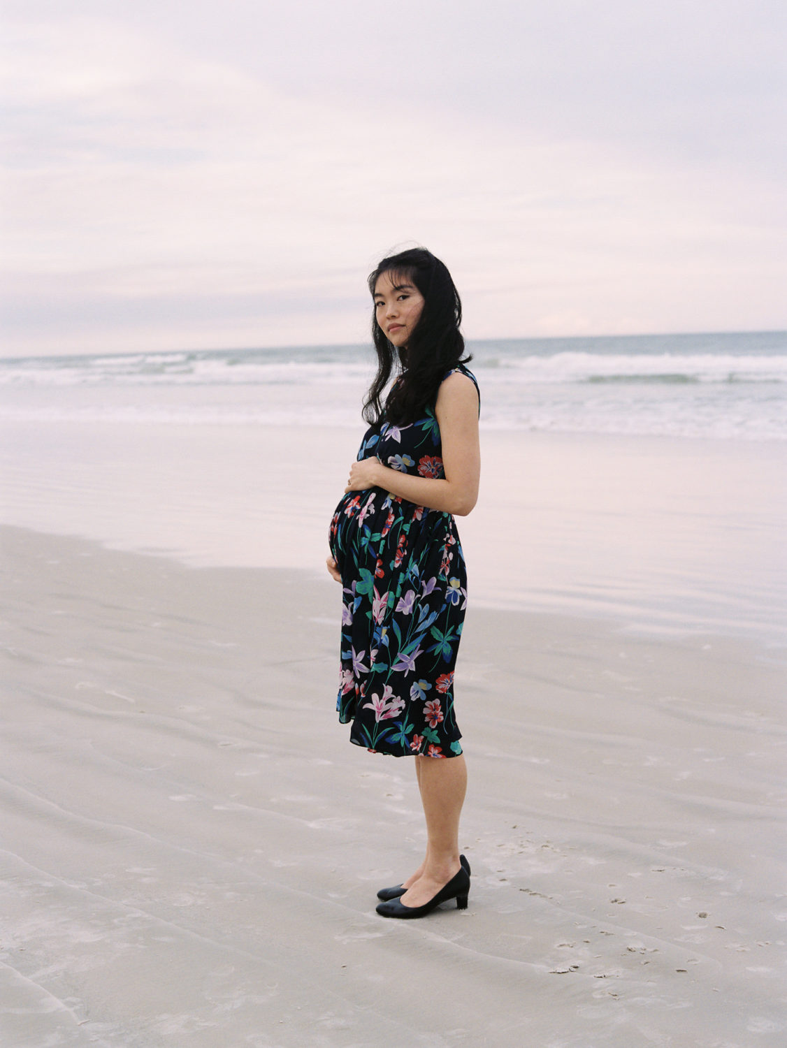 sideview of a korean american pregnant woman in a floral dress standing by the ocean at sunset