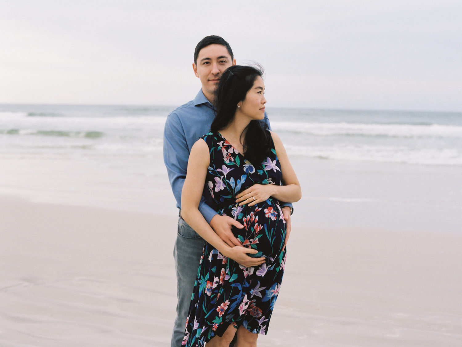 moody image of asian american husband and pregnant wife standing on the beach and looking seriously at the camera