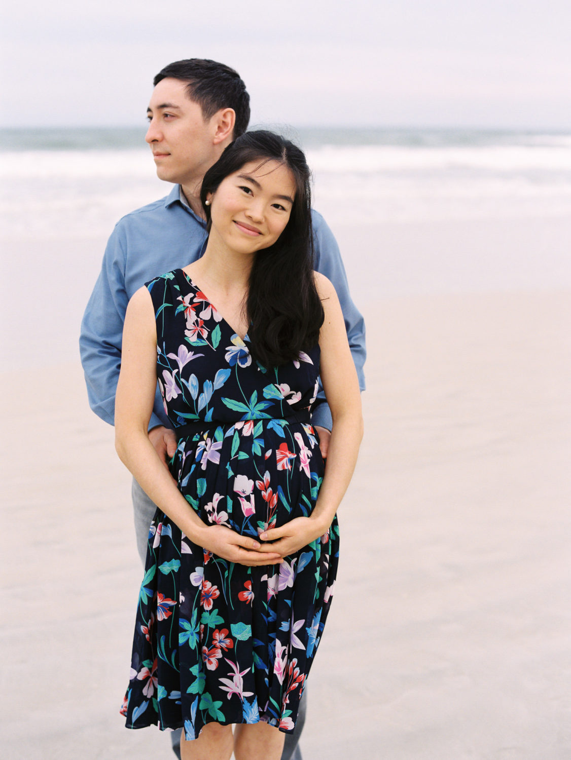 woman with long black hair and multicolored floral dress holding her hands under her pregnant belly and smiling