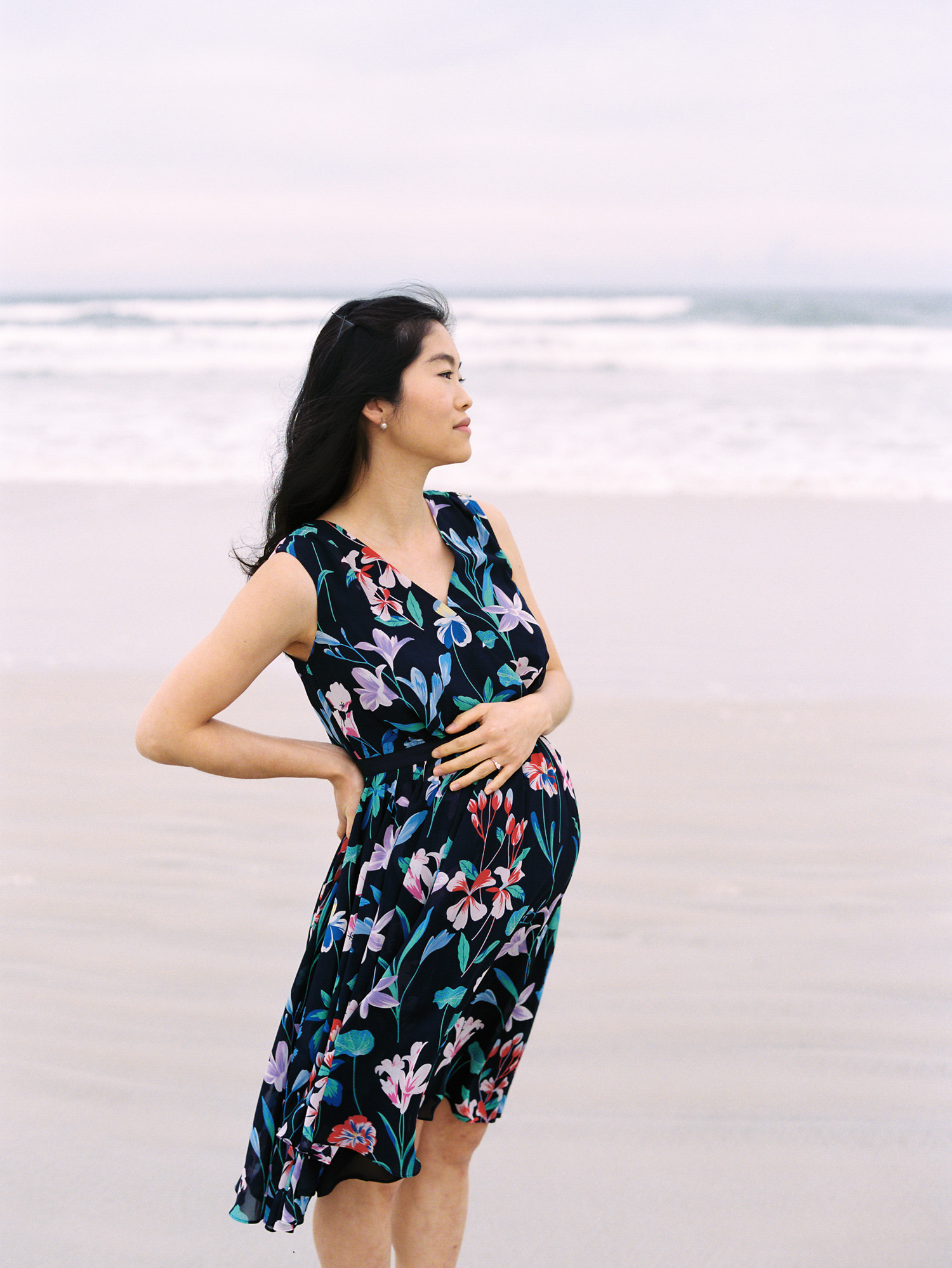 a pregnant korean woman standing on the beach with one hand on her back and the other resting on her pregnant belly during maternity photoshoot