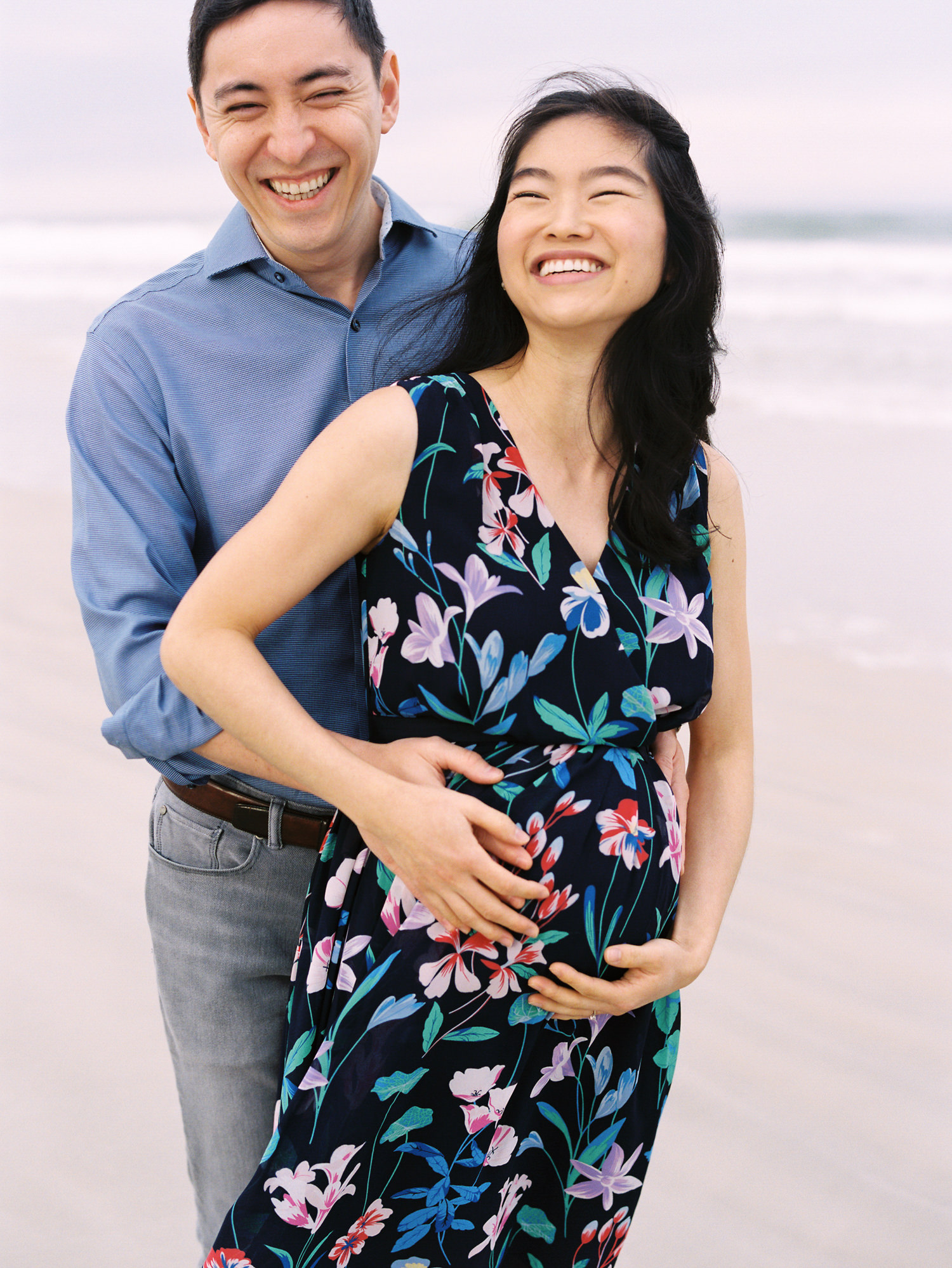 man in blue shirt standing behind pregnant wife in a blue floral dress putting their hands on her pregnant belly