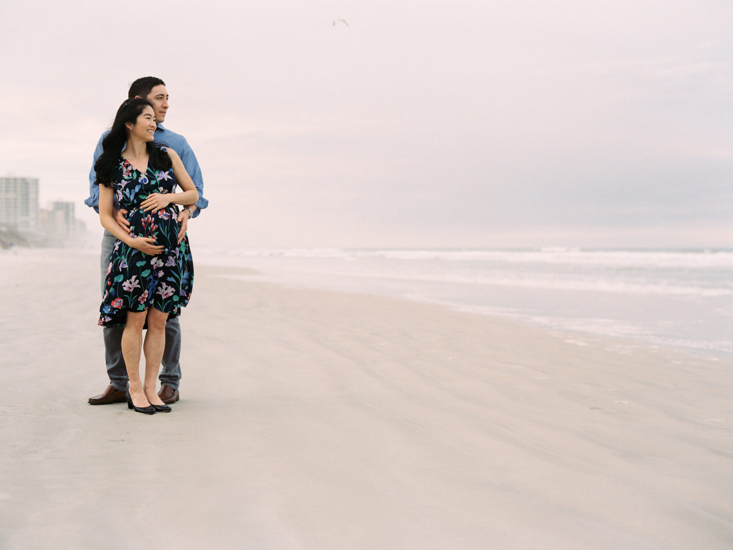 Man and his pregnant wife standing together on the beach smiling and looking at the ocean