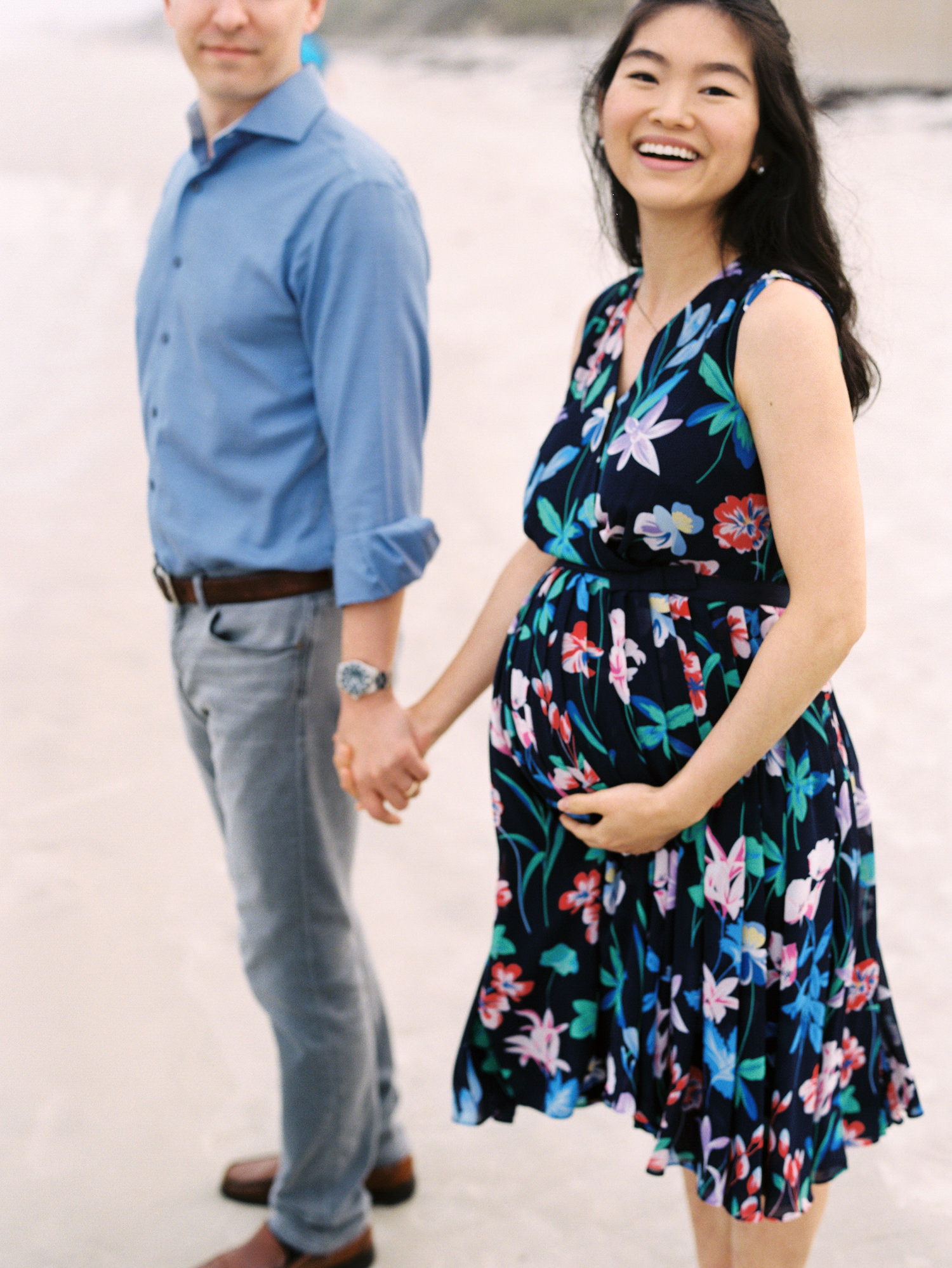 a pregnant woman in a multicolored floral dress holding her husband's hand and holding her baby bump with the other hand.