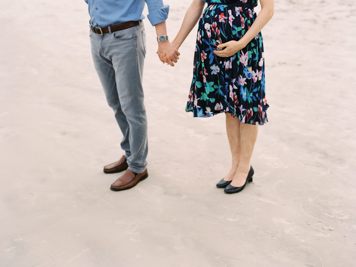 Close up of torso and legs of a man and pregnant woman holding hands and standing together on a sandy beach