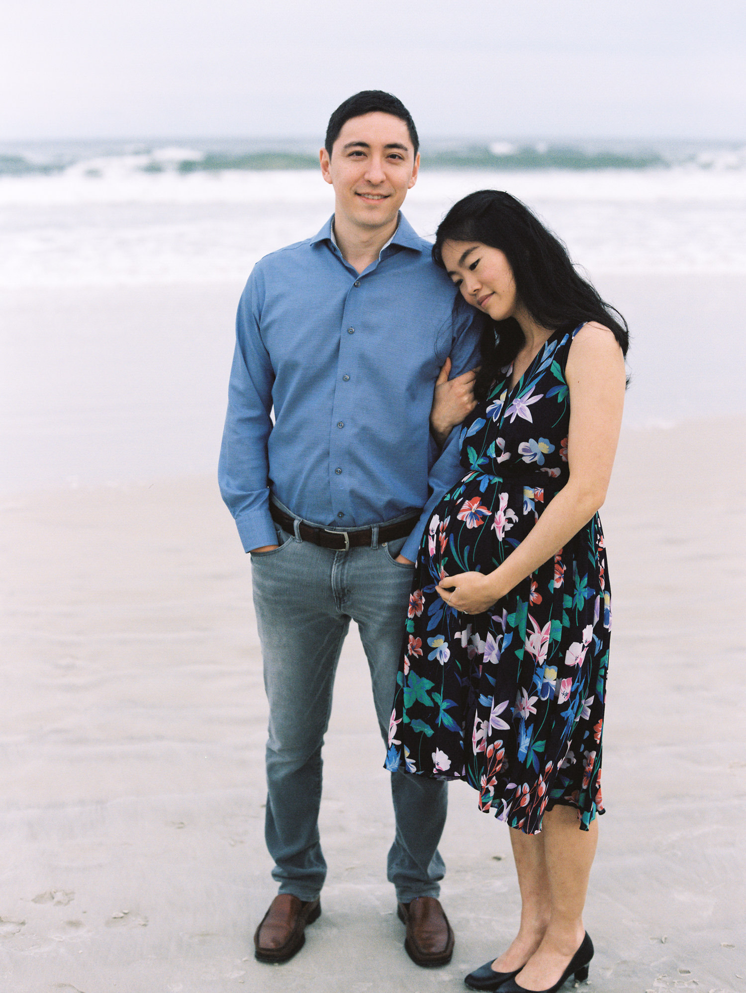 man with black hair and blue shirt and gray pants standing in front of ocean waves while his pregnant wife holds his arm and leans against his shoulder