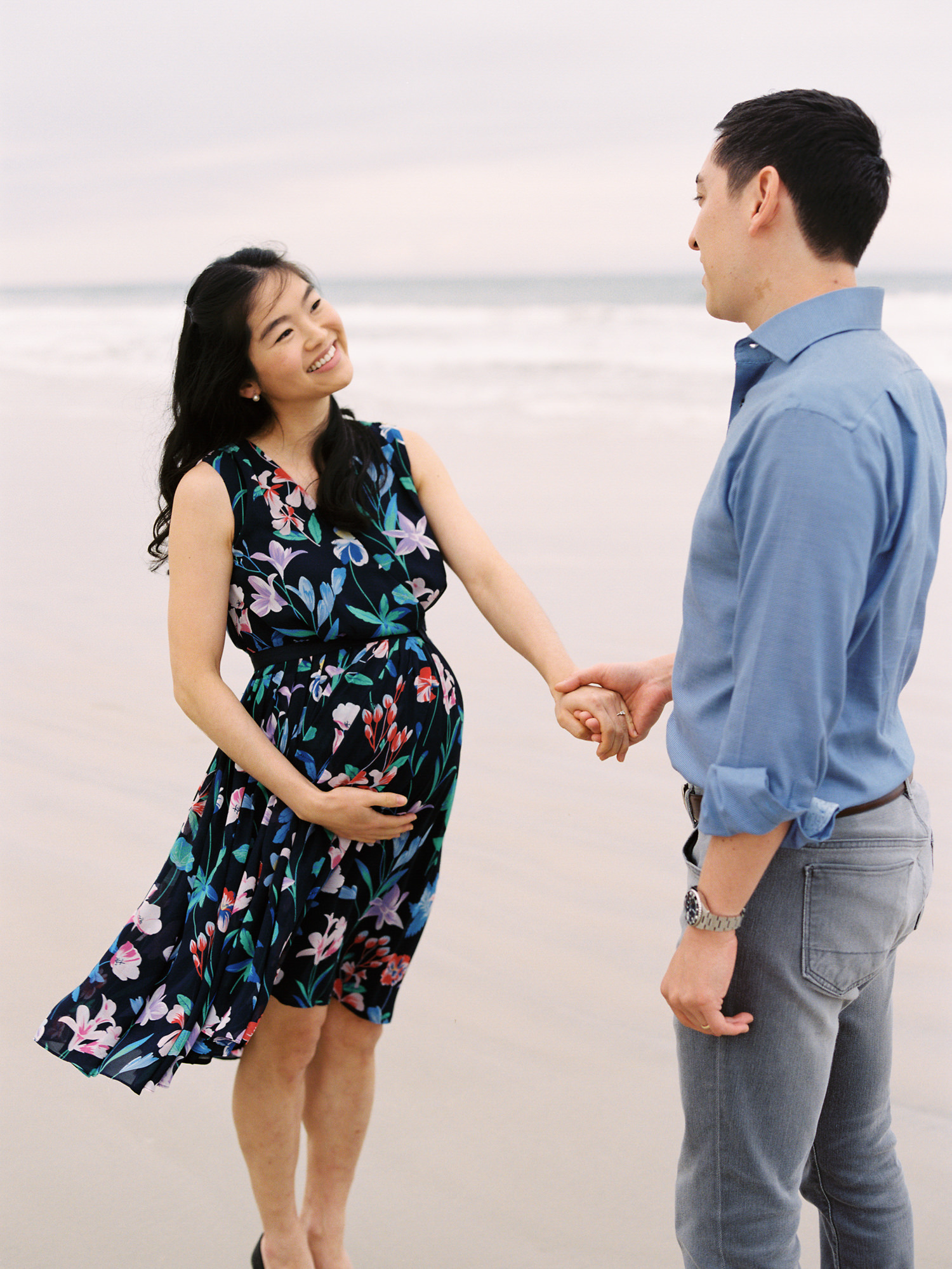 pregnant woman with long black hair and floral dress holding her husbands hand and smiling at him