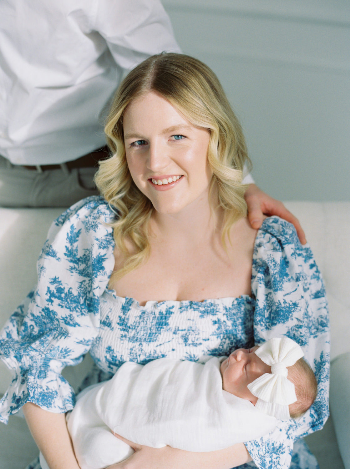 blonde blue eyed new mom wearing a blue and white dress smiling at the camera and holding her baby