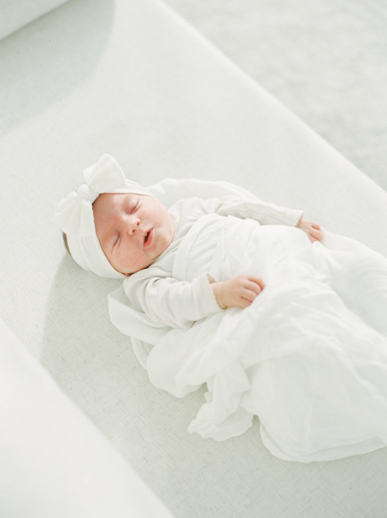 newborn baby girl in white onesie, white headband, and white blanket sleeping on a couch in the sun.