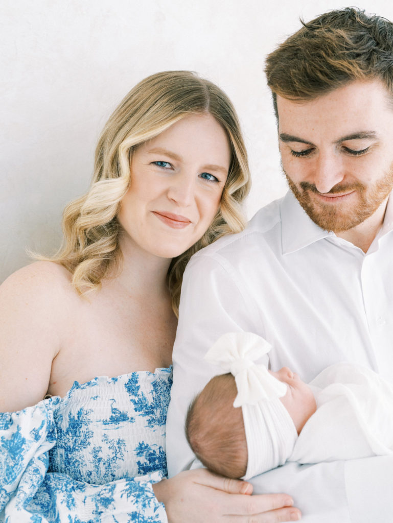 blonde new mom in blue and white dress smiling with her husband and new baby