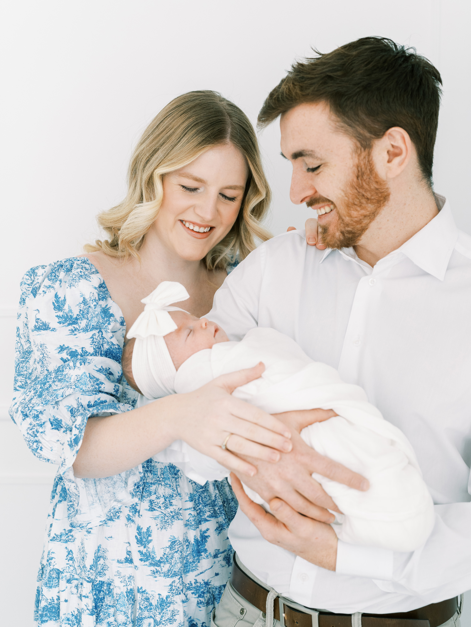 blonde new mother in blue and white dress standing next to her husband while he holds their new baby and smile down at them