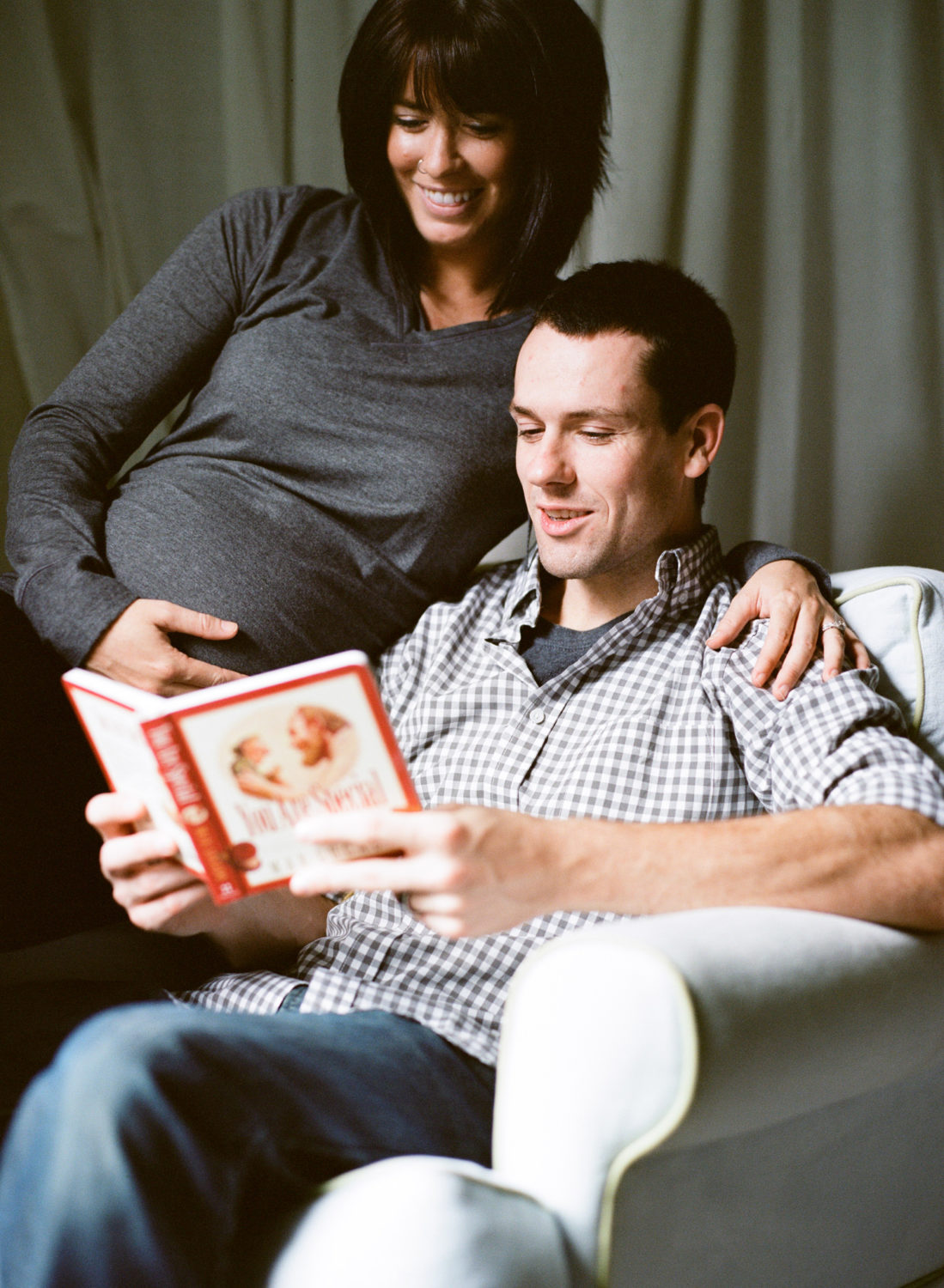 man reading a children's book as pregnant wife sits on the arm of the chair next to him